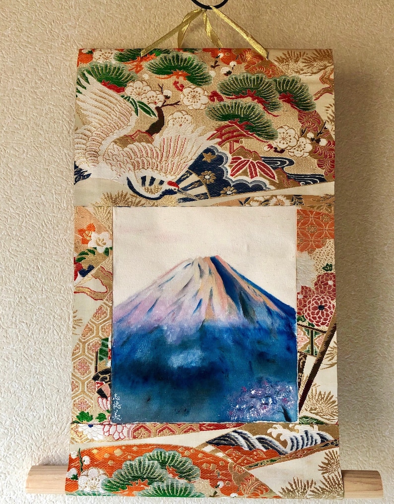 May include: A Japanese-style wall hanging featuring a painting of Mount Fuji with a snow-capped peak. The artwork is framed by ornate fabric with floral and crane patterns in gold, red, and orange. A wooden dowel is at the bottom.