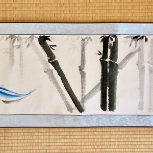 May include: A watercolor painting featuring bamboo stalks in shades of gray and black, with a blue fish swimming in the foreground. The artwork is framed with a light blue border and rests on a woven mat.