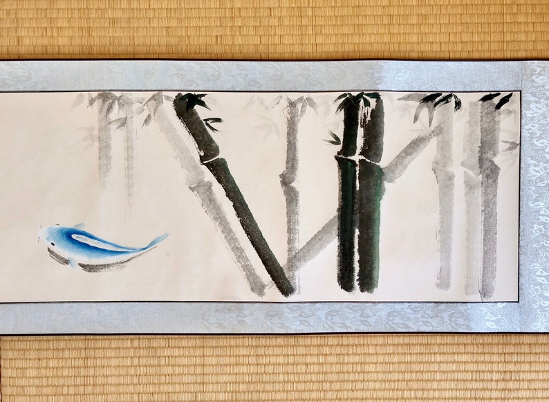 May include: A watercolor painting featuring bamboo stalks in shades of gray and black, with a blue fish swimming in the foreground. The artwork is framed with a light blue border and rests on a woven mat.