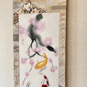 May include: A Japanese-style wall scroll featuring a watercolor painting of koi fish and cherry blossoms. The artwork includes a gold and white patterned fabric border and a wooden dowel at the bottom. A gold ribbon is attached at the top.