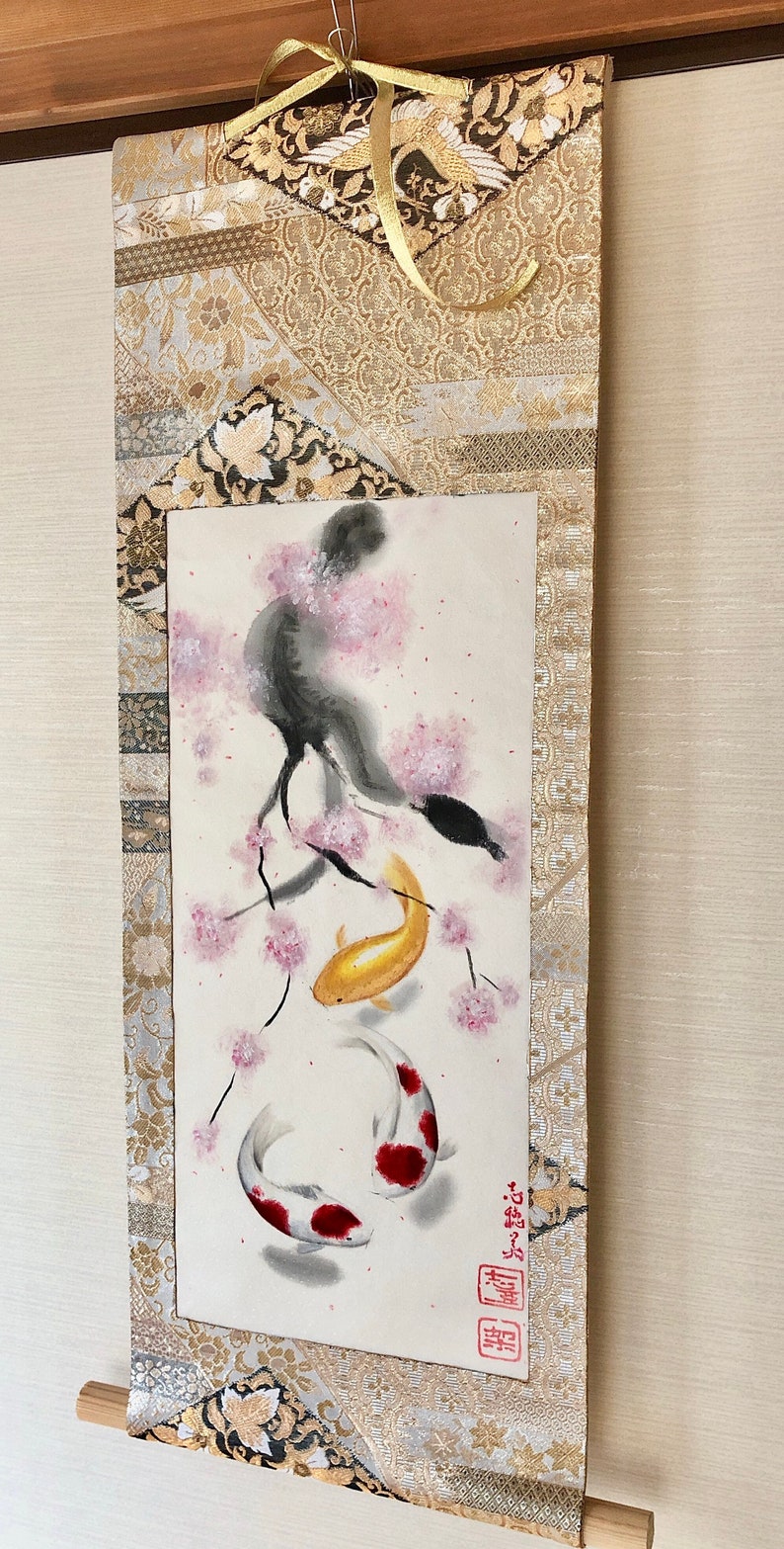 May include: A Japanese-style wall scroll featuring a watercolor painting of koi fish and cherry blossoms. The artwork includes a gold and white patterned fabric border and a wooden dowel at the bottom. A gold ribbon is attached at the top.