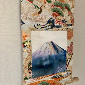 May include: Japanese wall scroll featuring a watercolor painting of Mount Fuji. The artwork is framed by a decorative fabric with gold accents, cranes, and floral patterns. A wooden dowel is at the bottom.