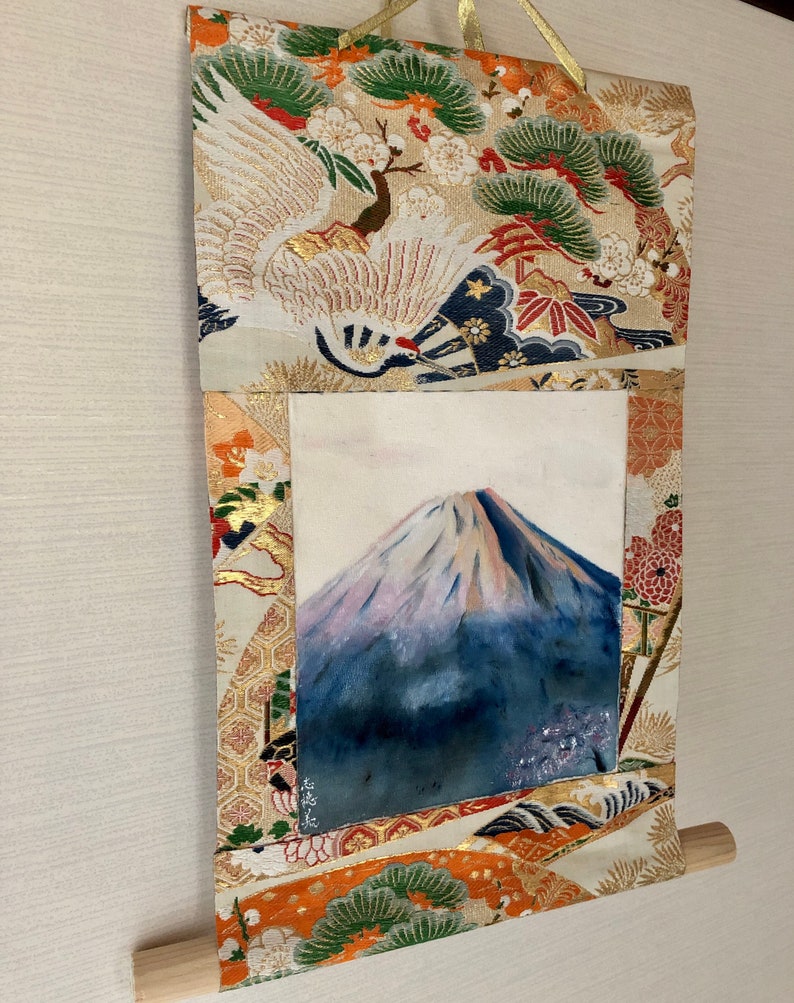 May include: Japanese wall scroll featuring a watercolor painting of Mount Fuji. The artwork is framed by a decorative fabric with gold accents, cranes, and floral patterns. A wooden dowel is at the bottom.