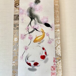 May include: Japanese-style scroll painting featuring two koi fish swimming beneath a flowering cherry blossom branch. The koi are gold and white with red markings. The scroll has a gold and beige patterned border.