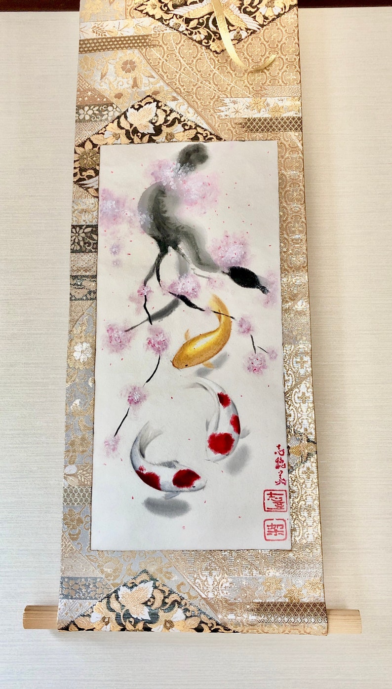 May include: Japanese-style scroll painting featuring two koi fish swimming beneath a flowering cherry blossom branch. The koi are gold and white with red markings. The scroll has a gold and beige patterned border.