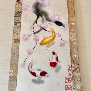 May include: Japanese-style wall scroll featuring a watercolor painting of koi fish and cherry blossoms. The artwork includes a gold koi, two white and red koi, and pink blossoms on a light background. The scroll is framed with gold and patterned fabric.
