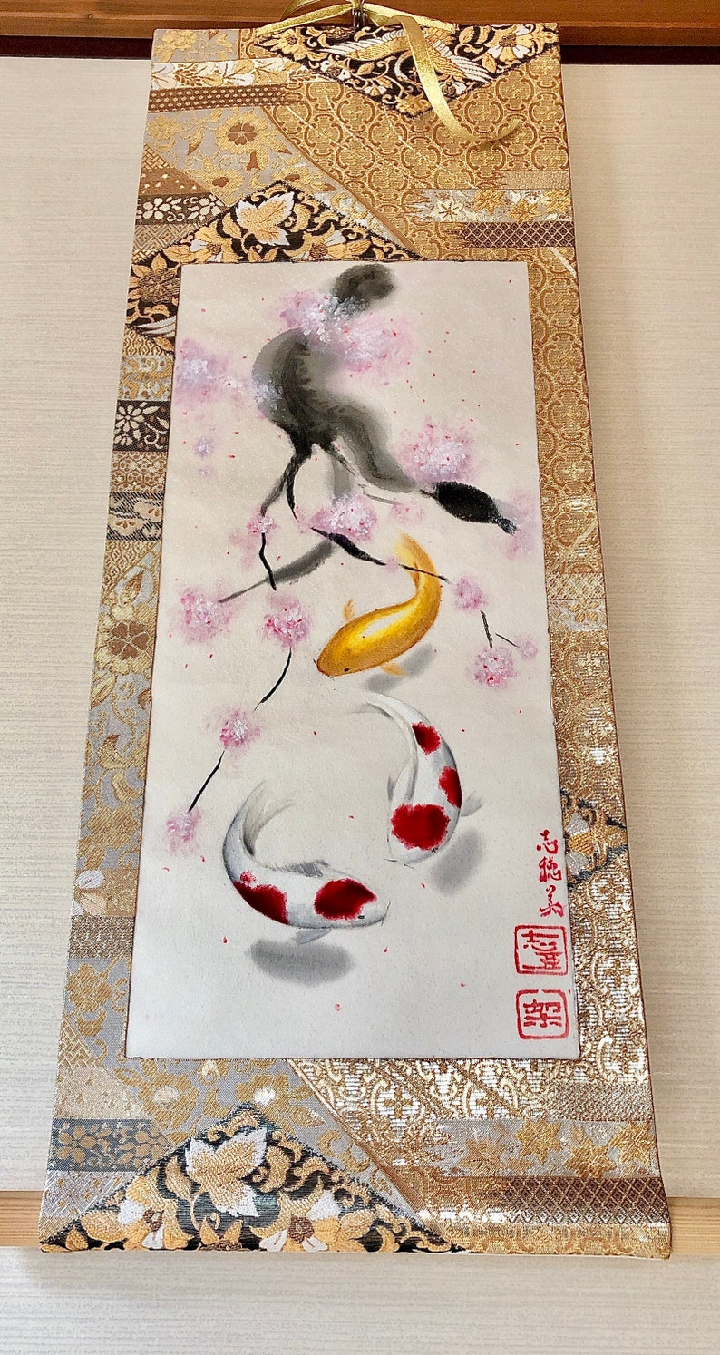 May include: Japanese-style wall scroll featuring a watercolor painting of koi fish and cherry blossoms. The artwork includes a gold koi, two white and red koi, and pink blossoms on a light background. The scroll is framed with gold and patterned fabric.