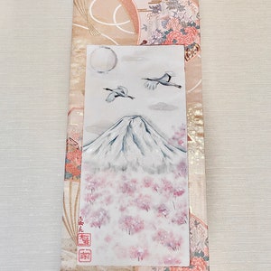 May include: Japanese wall scroll depicting Mount Fuji, cherry blossoms, cranes, and a full moon. The scroll has a beige and pink patterned border with floral and fan motifs. A wooden dowel is at the bottom.
