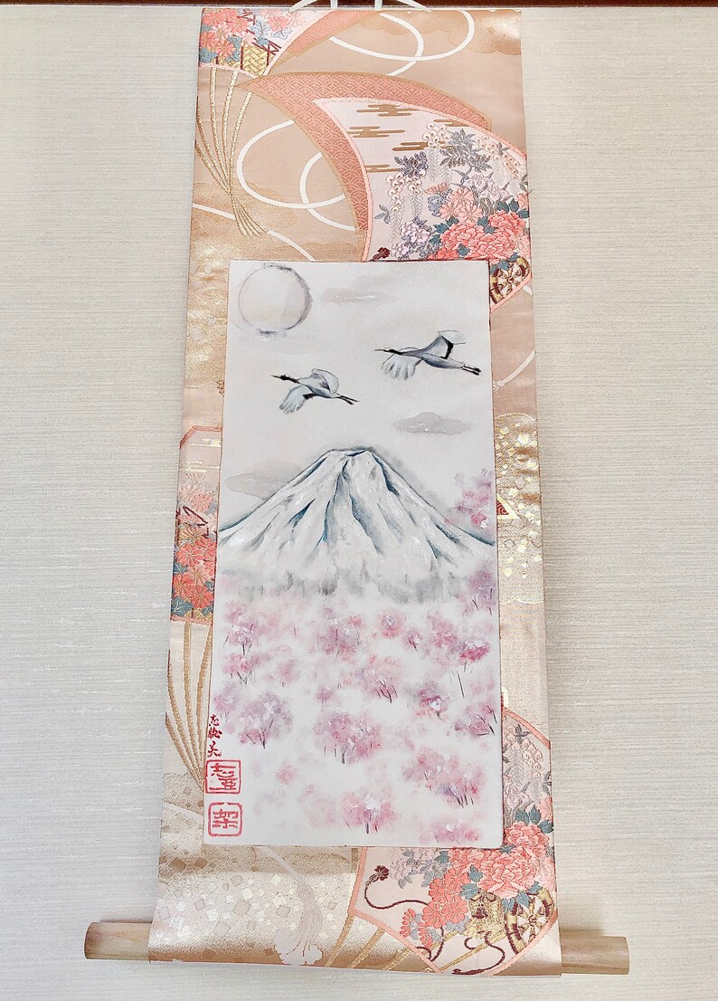 May include: Japanese wall scroll depicting Mount Fuji, cherry blossoms, cranes, and a full moon. The scroll has a beige and pink patterned border with floral and fan motifs. A wooden dowel is at the bottom.