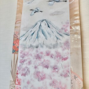 May include: An original painting of Mount Fuji with cherry blossom trees. Two cranes fly in a cloudy sky above the snow-covered mountain. The artwork features a full moon and is framed by decorative fabric.