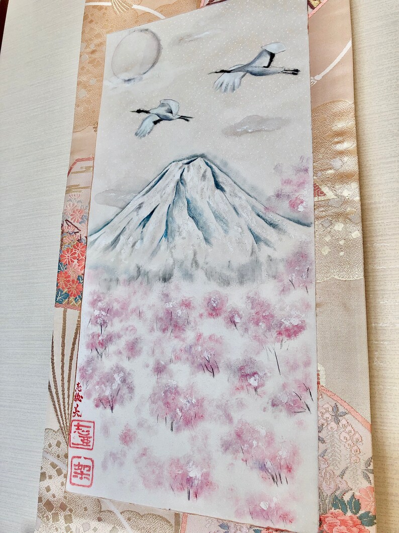 May include: An original painting of Mount Fuji with cherry blossom trees. Two cranes fly in a cloudy sky above the snow-covered mountain. The artwork features a full moon and is framed by decorative fabric.