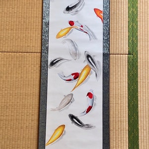 May include: A vertical scroll painting featuring koi fish in shades of red, orange, yellow, black, and white. The fish are depicted swimming in a minimalist style against a white background. The scroll has a dark green patterned border and wooden dowels.