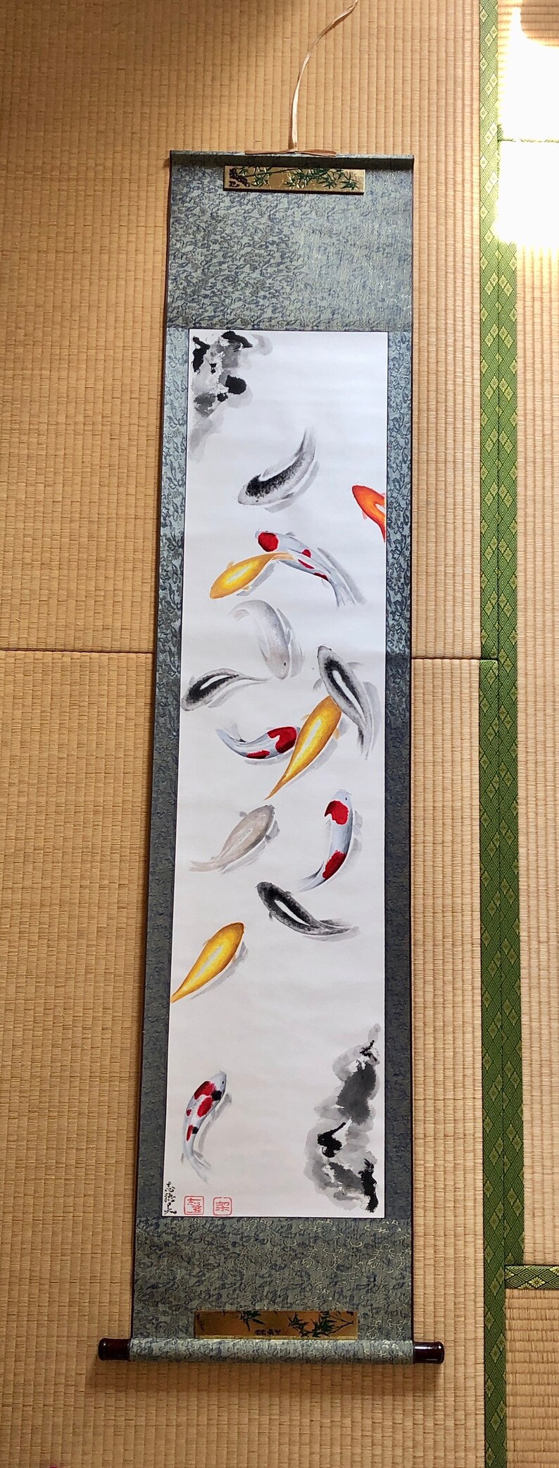 May include: A vertical scroll painting featuring koi fish in shades of red, orange, yellow, black, and white. The fish are depicted swimming in a minimalist style against a white background. The scroll has a dark green patterned border and wooden dowels.