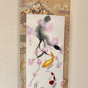 May include: Japanese scroll painting featuring a watercolor illustration of koi fish swimming beneath a flowering cherry blossom tree. The artwork is framed with gold brocade fabric and a wooden dowel at the bottom.