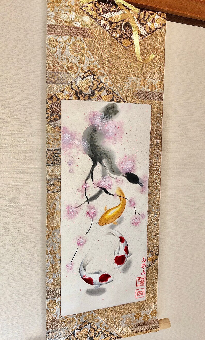 May include: Japanese scroll painting featuring a watercolor illustration of koi fish swimming beneath a flowering cherry blossom tree. The artwork is framed with gold brocade fabric and a wooden dowel at the bottom.