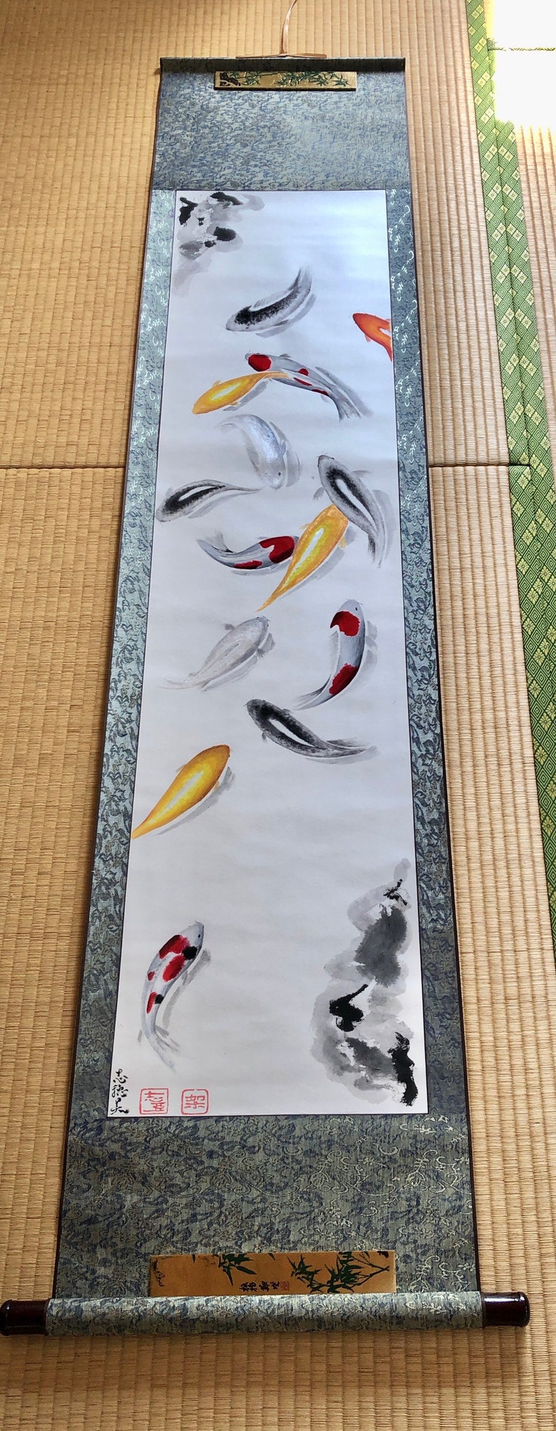 May include: A Japanese scroll painting featuring koi fish in shades of red, yellow, black, and white. The artwork is mounted on a dark blue fabric with a decorative border and wooden dowels.