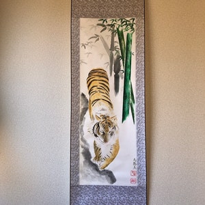 May include: A traditional Japanese scroll painting featuring a tiger with orange and black stripes, walking towards the viewer. The background includes bamboo stalks and foliage, with a decorative gray border.