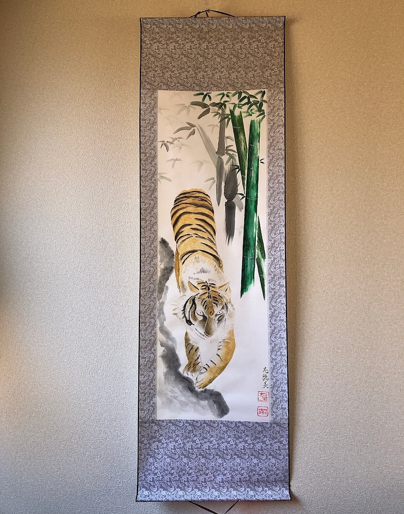 May include: A traditional Japanese scroll painting featuring a tiger with orange and black stripes, walking towards the viewer. The background includes bamboo stalks and foliage, with a decorative gray border.