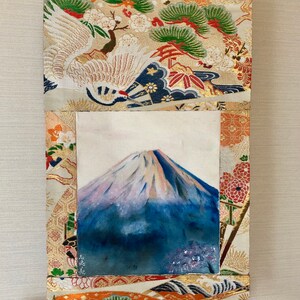 May include: Japanese-style wall scroll featuring a painting of Mount Fuji. The artwork is framed by a decorative fabric with crane and floral patterns in gold, orange, and green. A wooden dowel is at the bottom, and a gold cord is at the top.