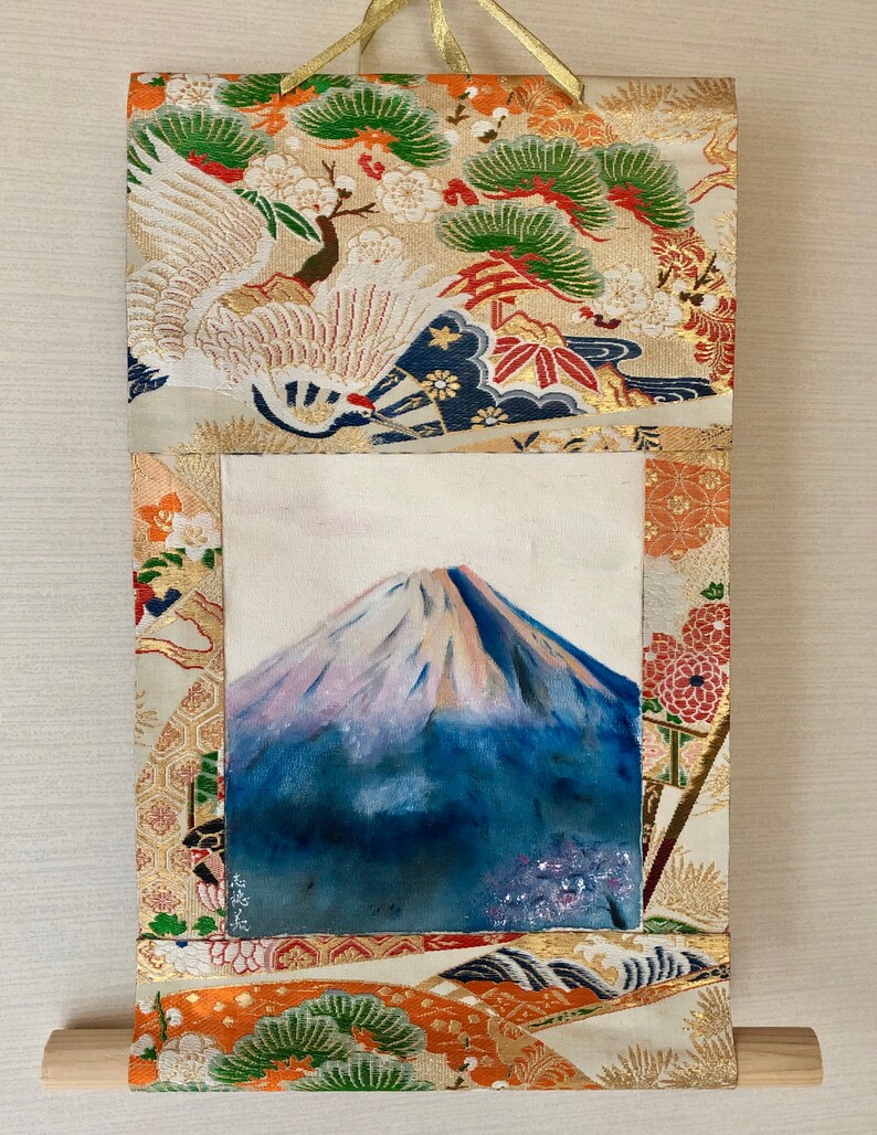May include: Japanese-style wall scroll featuring a painting of Mount Fuji. The artwork is framed by a decorative fabric with crane and floral patterns in gold, orange, and green. A wooden dowel is at the bottom, and a gold cord is at the top.