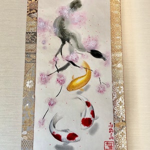 May include: Japanese scroll painting featuring two koi fish swimming beneath a cherry blossom branch. The koi are gold and white with red markings. The scroll has a gold and silver patterned border and a wooden dowel at the bottom.
