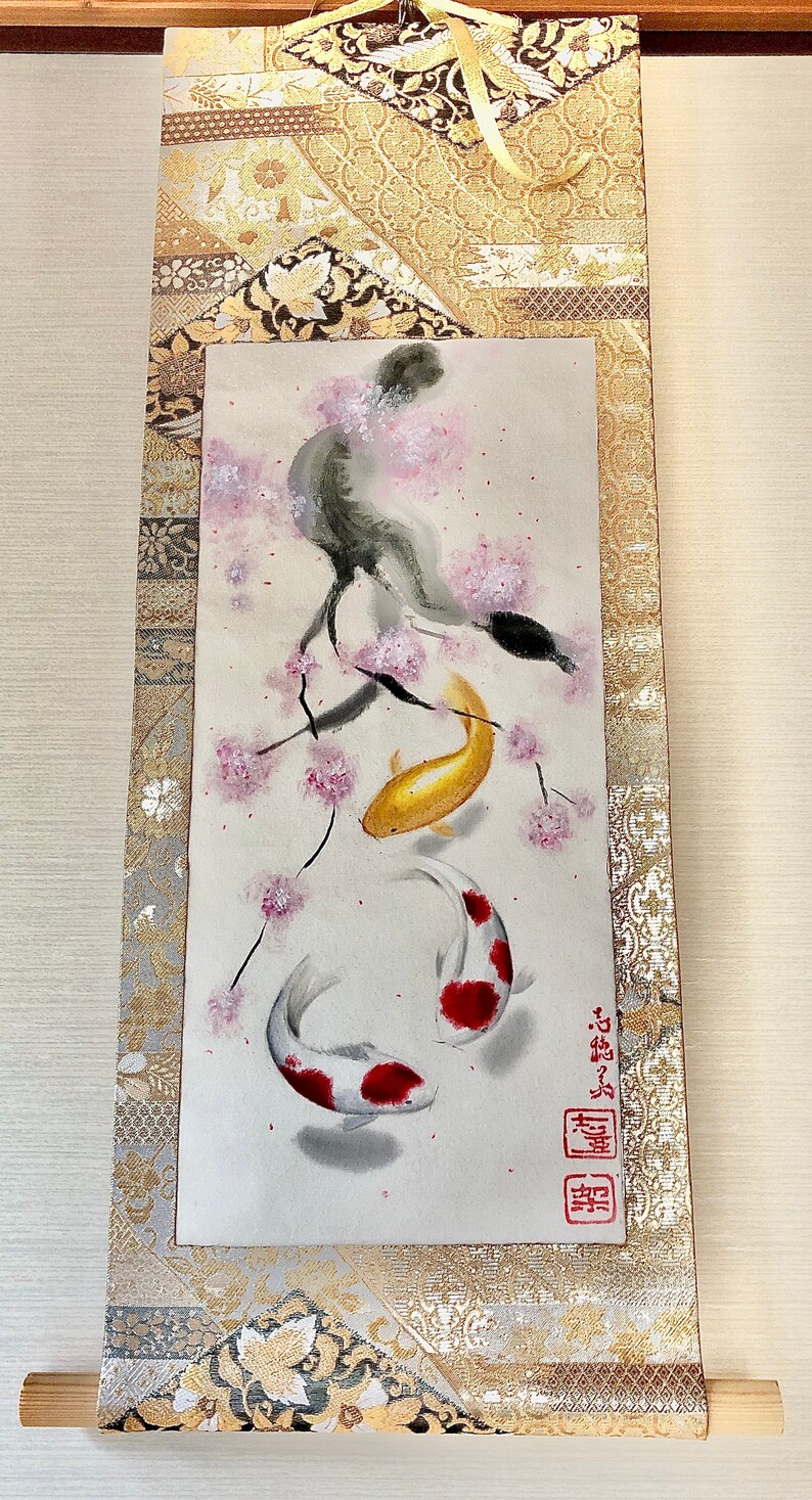 May include: Japanese scroll painting featuring two koi fish swimming beneath a cherry blossom branch. The koi are gold and white with red markings. The scroll has a gold and silver patterned border and a wooden dowel at the bottom.