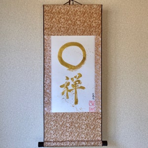 May include: A Japanese scroll painting featuring a gold Enso circle and calligraphy. The artwork includes the Japanese character for Zen, with gold paint splatters and red seals. The scroll has a brown and cream patterned border.