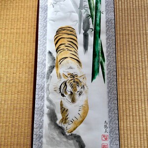 May include: A traditional Japanese scroll painting featuring a tiger with orange and black stripes, walking towards the viewer. The background includes bamboo stalks and foliage, with a gray and white patterned border.
