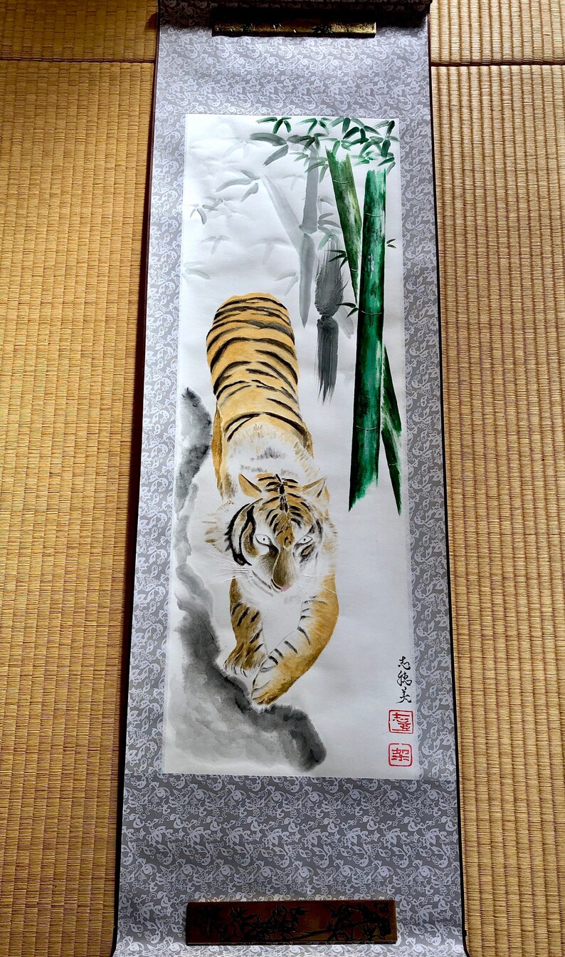 May include: A traditional Japanese scroll painting featuring a tiger with orange and black stripes, walking towards the viewer. The background includes bamboo stalks and foliage, with a gray and white patterned border.