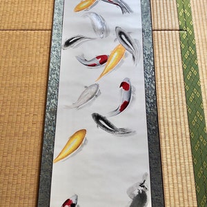 May include: A vertical scroll painting featuring koi fish in shades of black, white, red, and gold. The fish are depicted swimming in a flowing, abstract style. The artwork is framed with a decorative border and includes Japanese characters.