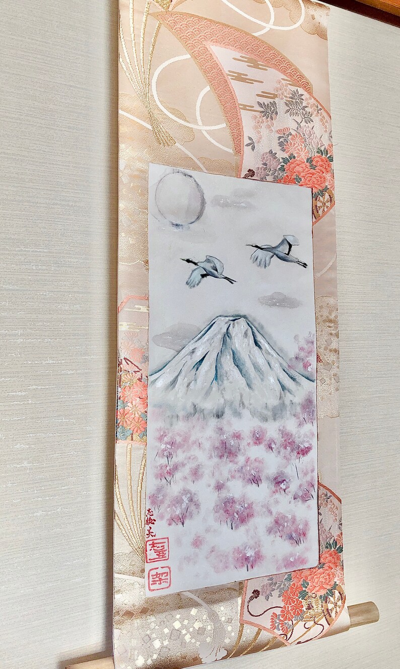 May include: Japanese scroll painting depicting Mount Fuji, cherry blossoms, and cranes. The artwork is set against a decorative fabric with floral and geometric patterns. The painting includes a full moon and clouds.