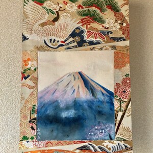 May include: A Japanese wall scroll featuring a painting of Mount Fuji with a blue and white color palette. The scroll is framed by ornate fabric with crane, floral, and wave patterns in gold, orange, and green. A wooden dowel is at the bottom.