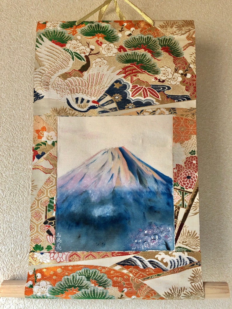 May include: A Japanese wall scroll featuring a painting of Mount Fuji with a blue and white color palette. The scroll is framed by ornate fabric with crane, floral, and wave patterns in gold, orange, and green. A wooden dowel is at the bottom.