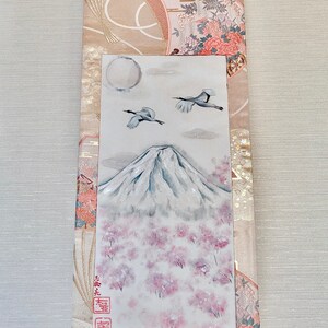 May include: A Japanese scroll painting depicting Mount Fuji with cherry blossom trees, two cranes, and a full moon. The artwork is set against a backdrop of patterned fabric in beige and pink tones. The scroll has a wooden dowel at the bottom.
