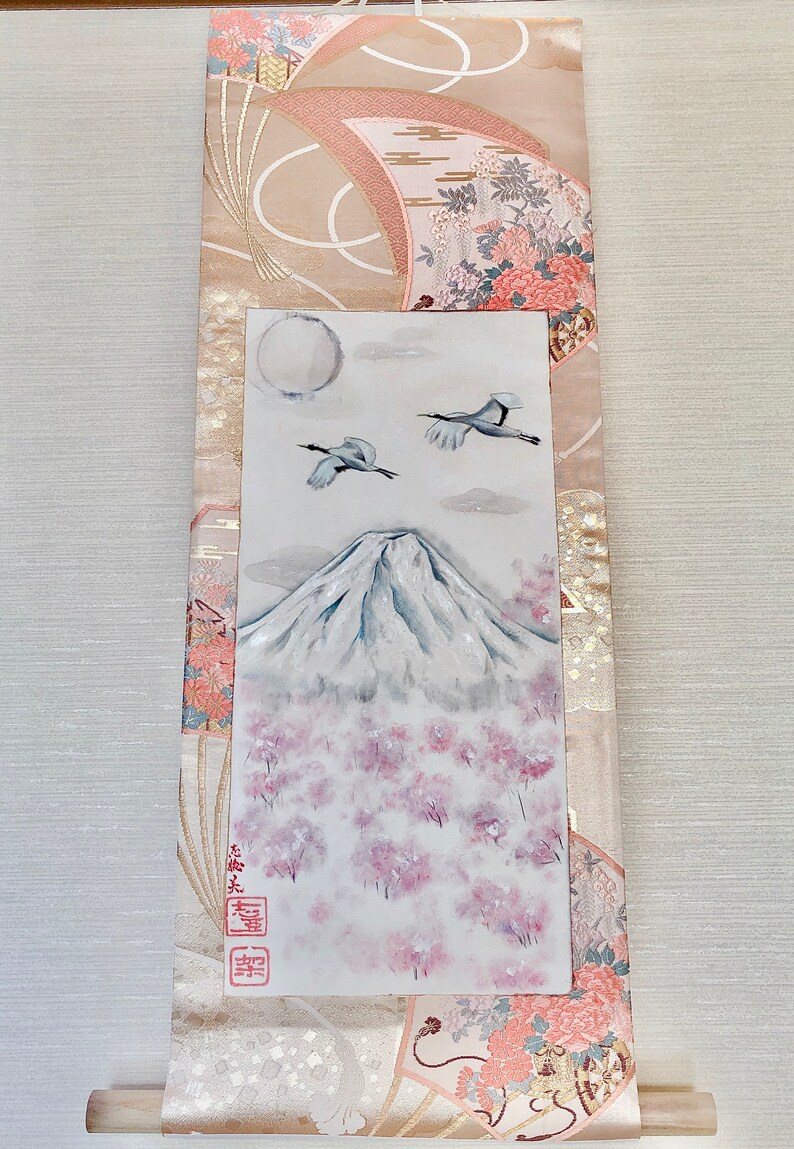 May include: A Japanese scroll painting depicting Mount Fuji with cherry blossom trees, two cranes, and a full moon. The artwork is set against a backdrop of patterned fabric in beige and pink tones. The scroll has a wooden dowel at the bottom.