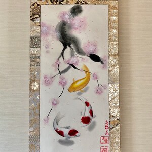 May include: Japanese scroll painting featuring two koi fish swimming beneath a cherry blossom branch. The artwork is in watercolor, with gold, red, and white accents. The scroll has a gold and black patterned border and a wooden dowel at the bottom.
