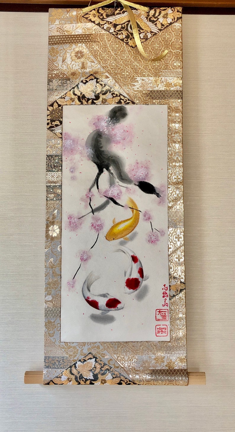 May include: Japanese scroll painting featuring two koi fish swimming beneath a cherry blossom branch. The artwork is in watercolor, with gold, red, and white accents. The scroll has a gold and black patterned border and a wooden dowel at the bottom.
