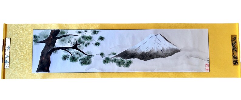 May include: A Japanese-style scroll painting featuring a mountain landscape. The artwork depicts a snow-capped mountain, a tree with green foliage, and a misty atmosphere. The scroll has a gold border and decorative end pieces.