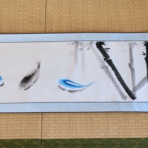 May include: A long, rectangular scroll painting featuring three fish and bamboo stalks. The fish are blue and gray, and the bamboo is rendered in black and gray. The scroll has a light blue border and wooden accents.