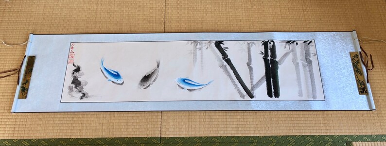 May include: A long, rectangular scroll painting featuring three fish and bamboo stalks. The fish are blue and gray, and the bamboo is rendered in black and gray. The scroll has a light blue border and wooden accents.