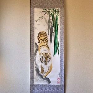 May include: A traditional Japanese scroll painting featuring a tiger with orange and black stripes, walking towards the viewer. The artwork includes bamboo stalks and foliage, with a gray patterned border.