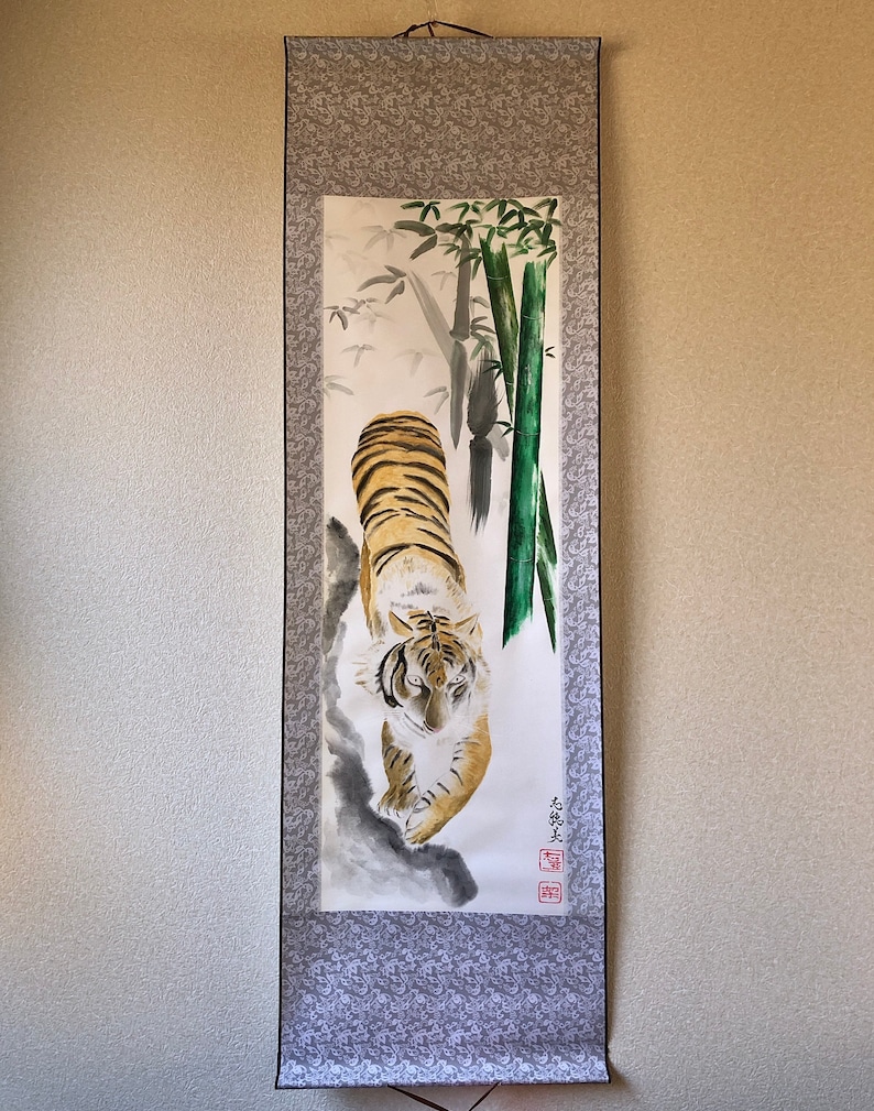 May include: A traditional Japanese scroll painting featuring a tiger with orange and black stripes, walking towards the viewer. The artwork includes bamboo stalks and foliage, with a gray patterned border.