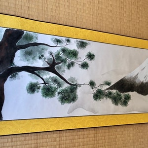 May include: A Japanese-style scroll painting featuring a tree and a mountain. The tree is dark brown with green foliage, and the mountain is gray with a white peak. The scroll has a gold border and is displayed on a tatami mat.