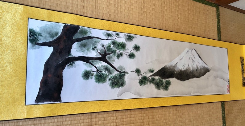 May include: A Japanese-style scroll painting featuring a tree and a mountain. The tree is dark brown with green foliage, and the mountain is gray with a white peak. The scroll has a gold border and is displayed on a tatami mat.