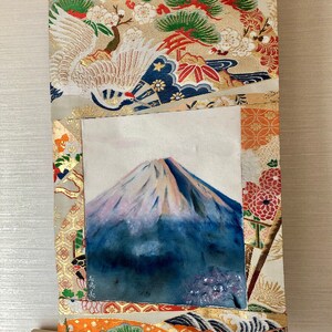 May include: A Japanese-style wall scroll featuring a painting of Mount Fuji with a snow-capped peak. The scroll is framed with ornate fabric in gold, orange, and cream colors, with traditional Japanese motifs including cranes and pine trees. A wooden dowel is at the bottom.