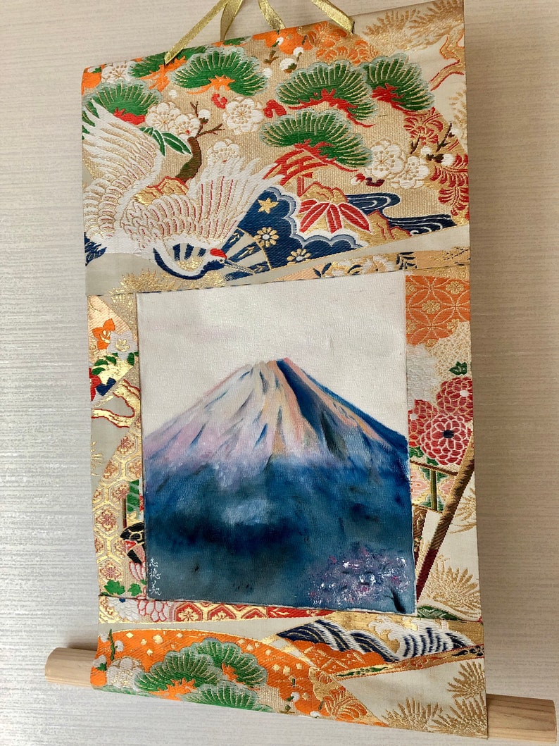 May include: A Japanese-style wall scroll featuring a painting of Mount Fuji with a snow-capped peak. The scroll is framed with ornate fabric in gold, orange, and cream colors, with traditional Japanese motifs including cranes and pine trees. A wooden dowel is at the bottom.