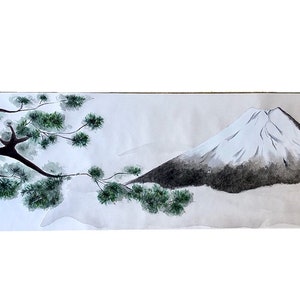 May include: Watercolor painting of Mount Fuji with a snow-capped peak, a dark tree trunk, and green foliage. The artwork is on a long, rectangular piece of paper, with Japanese characters in the lower right corner.