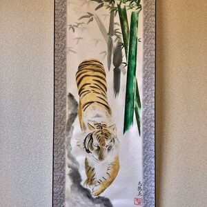 May include: A traditional Japanese scroll painting featuring a tiger with orange and black stripes, walking towards the viewer. The background includes bamboo stalks and foliage in shades of green and gray. The scroll is framed with a patterned border.