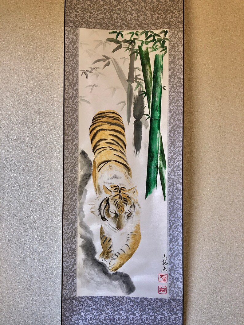 May include: A traditional Japanese scroll painting featuring a tiger with orange and black stripes, walking towards the viewer. The background includes bamboo stalks and foliage in shades of green and gray. The scroll is framed with a patterned border.