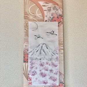 May include: Japanese wall scroll depicting Mount Fuji, cherry blossoms, cranes, and a full moon. The scroll has a beige and pink patterned fabric border with floral and fan motifs. A wooden dowel is at the bottom.